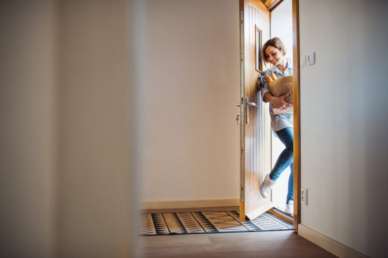 A young woman with groceries in paper shopping bag walking in through front door at home.